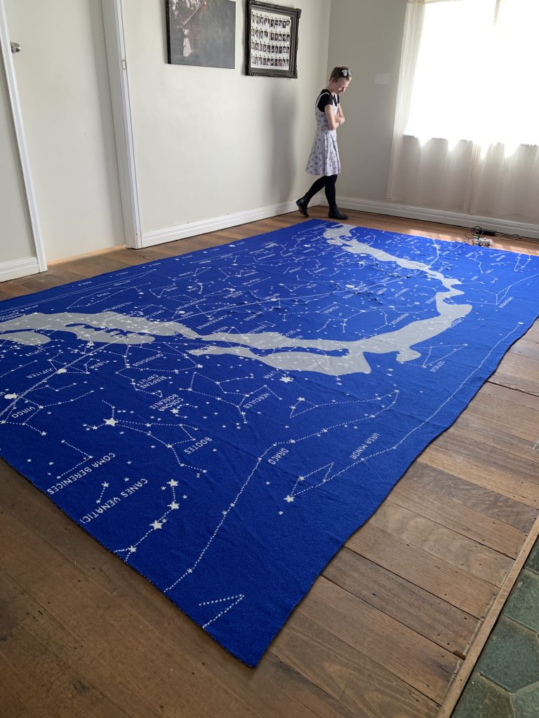 A person looks down at a large, knitted map of the stars on a wooden floor. The map is largely knitted in blue wool.