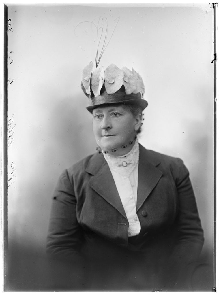 photographic portrait of woman wearing a decorative hat with flowers attached, a net face covering and a cameo broach