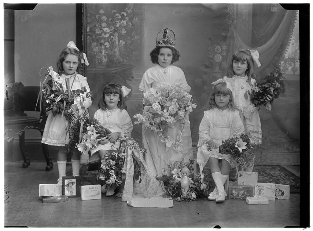 photographic portrait of 5 young children dressed in white carrying bouquets of flowers, one girl in the centre wearing a crown