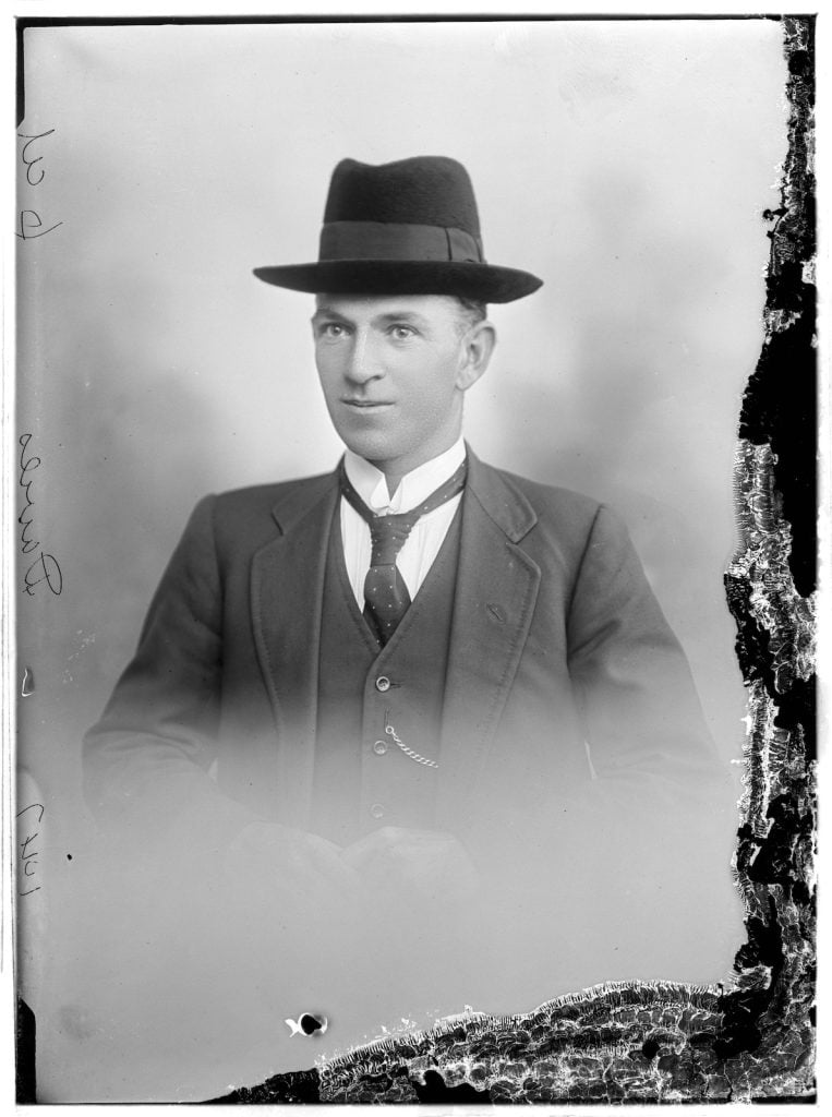 photographic portrait of a young man wearing a suit with a wing collared shirt, and a hat.