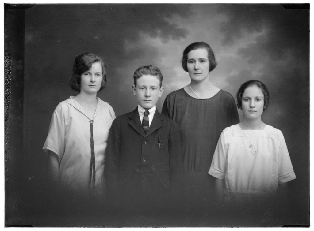 photographic portrait of a woman with 3 children, a family grouping. Woman wearing black and one girl in a white dress with a sailor typecollar, the boy in a jacket with a pen in the top pocket, the girl in a white dress with a square neckline.
