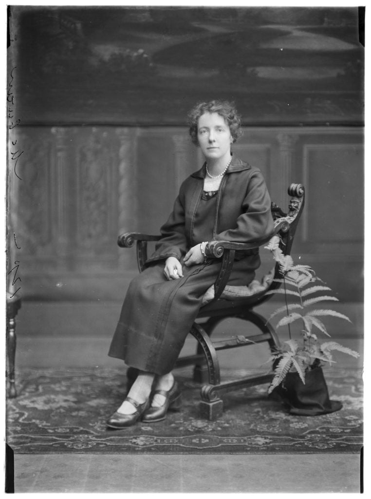photographic portrait of woman sitting on a chair in a black suit, with a string of pearls, a potted fern beside the chair