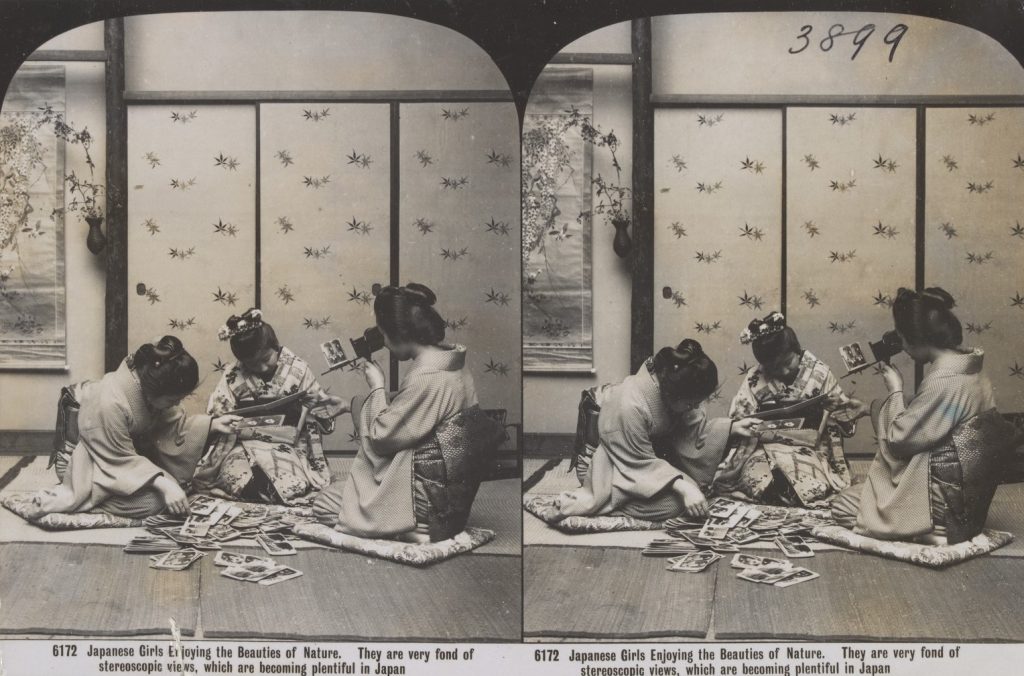 Three Japanese girls wearing kimonos sit on floor and view stereograph photographs. One girl views a stereograph through a stereoscope. 