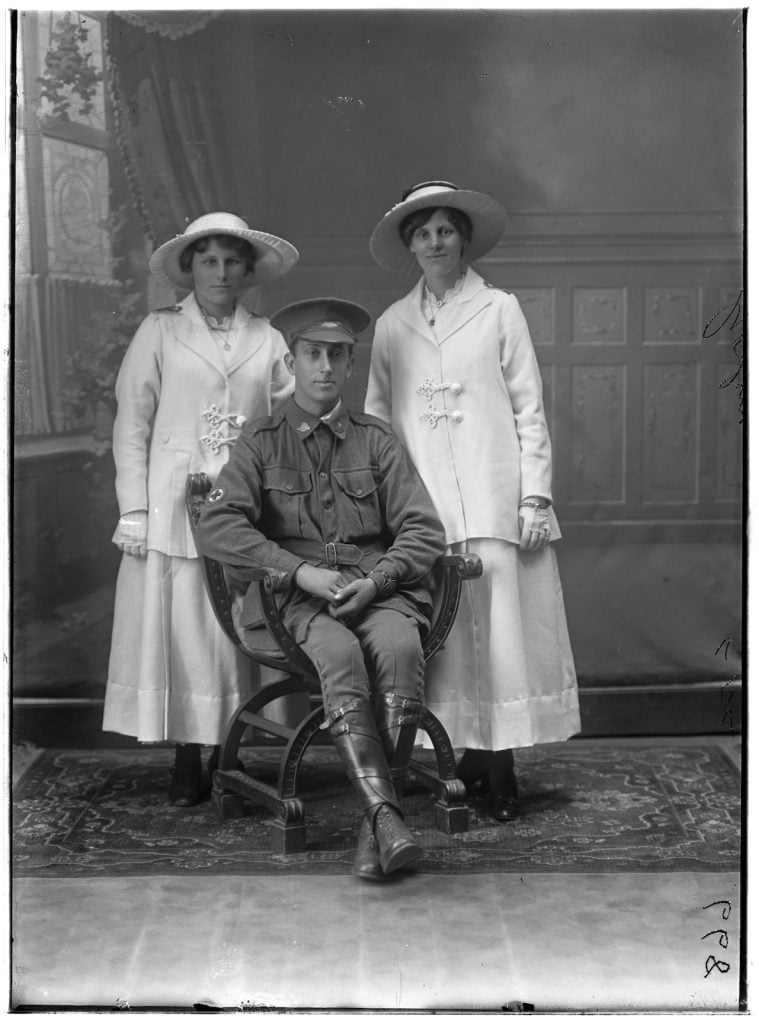 two women, in nurses uniforms - white jackets and white wide brimmed hats, one with a red cross badge at her neck. A seated soldier with a red cross patch on his sleeve