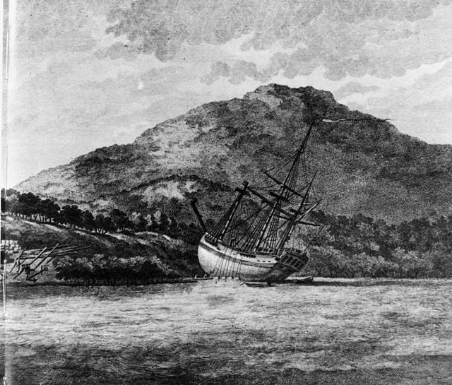 Black and white illustration of Captain Cook's ship Endeavour being careened on an Australian shore, at the foot of a hill.