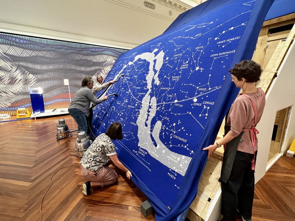 Using their hands, several people support a large frame with a blue knitted tapestry of the stars covering it. They hold in an upright position, while two others are vacuuming the centre area of the artwork. 