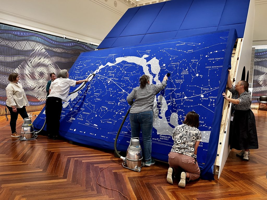Using their hands, three people support a large frame with a blue knitted tapestry of the stars covering it. They hold the frame in an upright position while two others are vacuuming the centre of the artwork. One person stands slightly apart, watching the vacuuming taking place. 