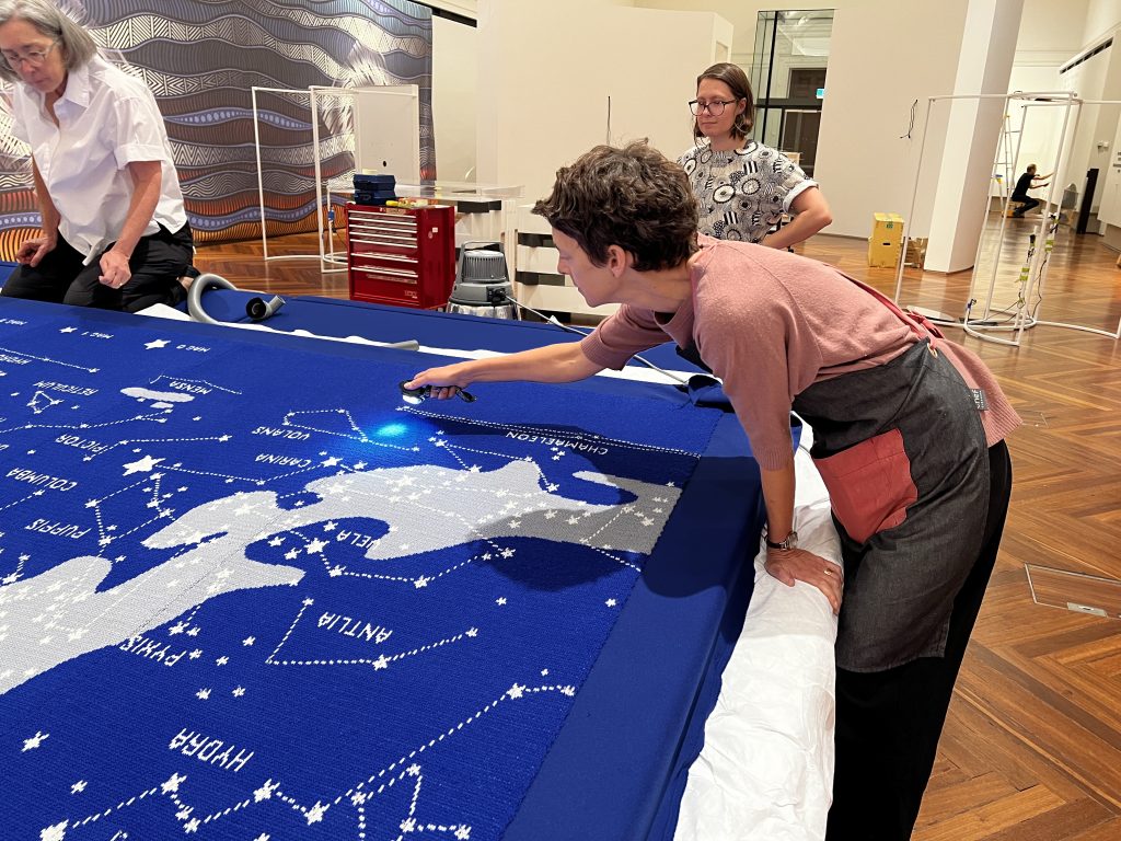 A person bends across the edge of a large blue knitted tapestry of the stars, holding a torch in one hand close to the tapestry. A blue circle of light falls on the tapestry below the torch. Two other people look on in the background.