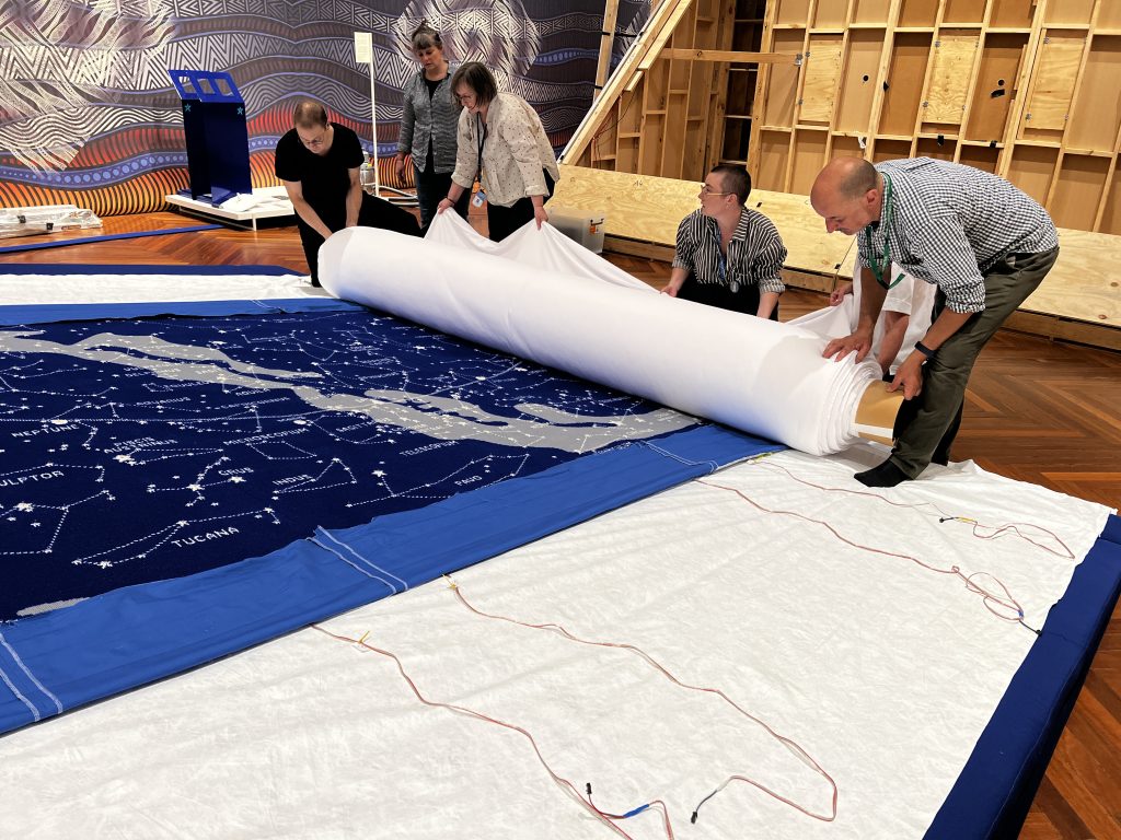 Four people are bent forward, mid-way through unrolling a large roll of white fabric across a blue knitted tapestry of the stars. The tapestry is laying horizontally on a large seat shaped like a wedge. One person looks on in the background.