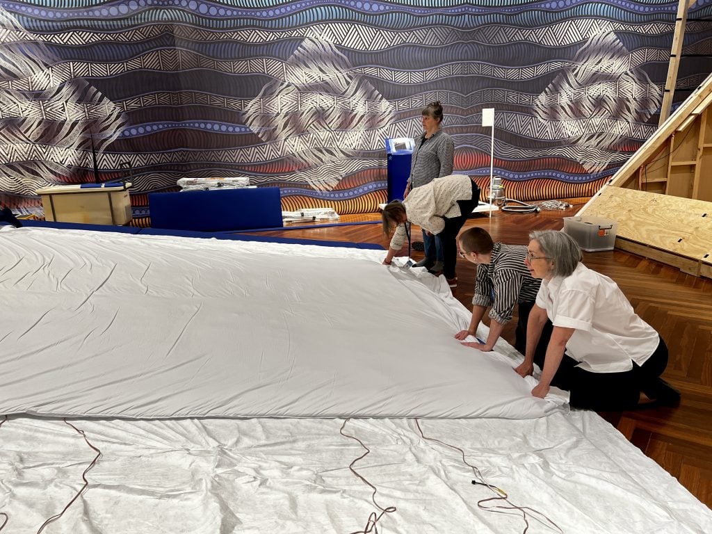 Three people are bent forward, holding tight to the short edge of a large piece of white fabric. The fabric rests horizontally on a large seat shaped like a wedge. One person looks on in the background.