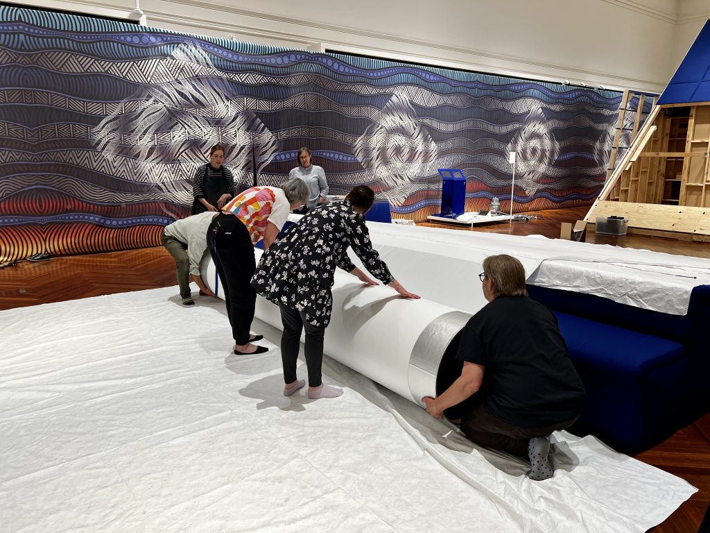 Two people are bent towards a large tube covered in silver foil. Two people at either end of the tube are knelt down in preparation to lift the tube up off the floor. The tube has a piece of white fabric wrapped around it. Two people in the background look on. 