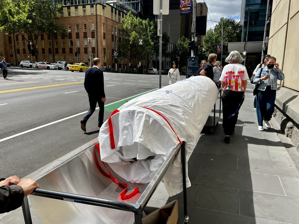 View of a large cylindrical object wrapped in white material guided along a footpath, its ends supported atop two curved metal trolleys on wheels. A road is visible at left. 