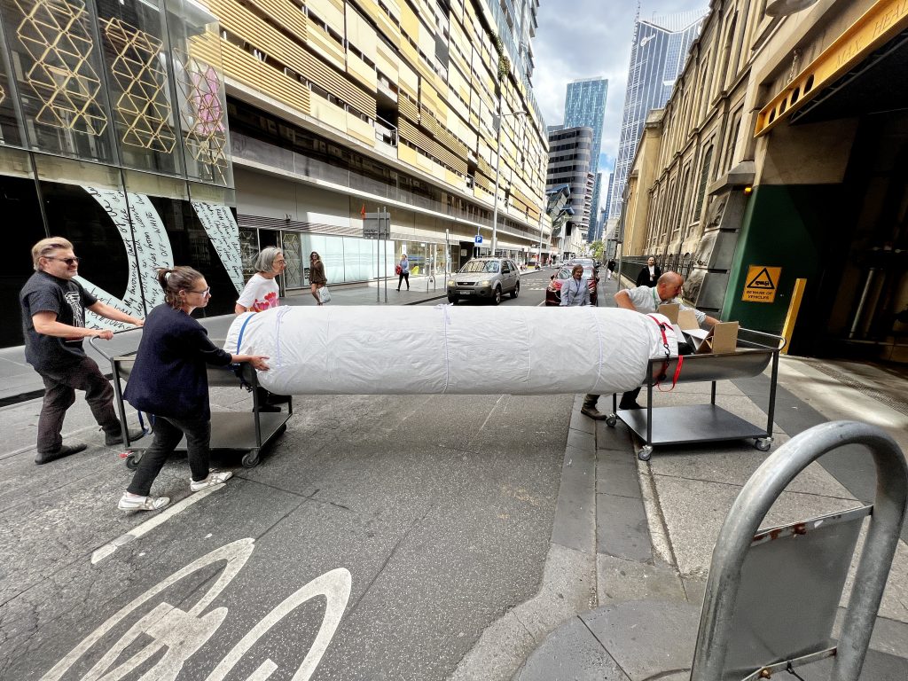 View of a large cylindrical object wrapped in white material being manoeuvred toward a loading dock across a road. The cylindrical object is supported by two curved metal trolleys on wheels, at both ends. Several people at either end of the cylindrical object push the trolleys toward the loading dock entrance. 