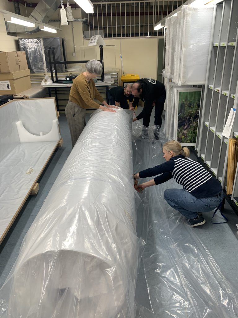Four people bend toward one end of a large cylindrical object wrapped in plastic, that rests on the floor. The room looks to be a storage space. 