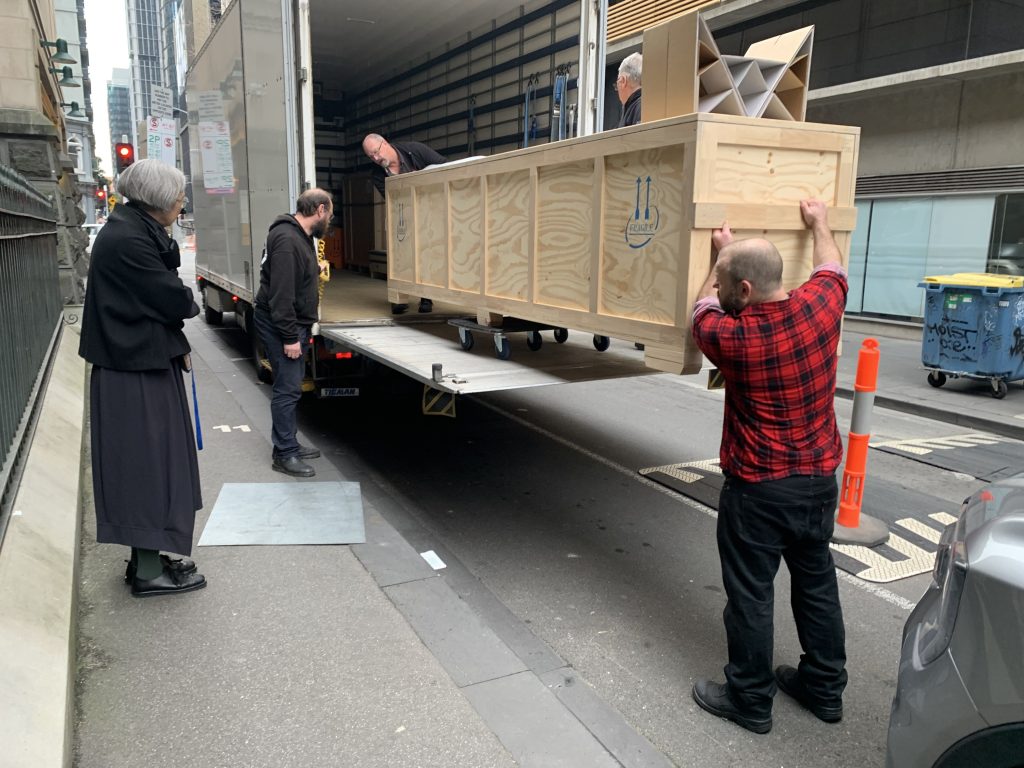 View of a large, long wooden crate elevated in the air on a platform attached to the back of a truck parked on a road. Three people are manoeuvring the crate into the truck. Two other people look on. 