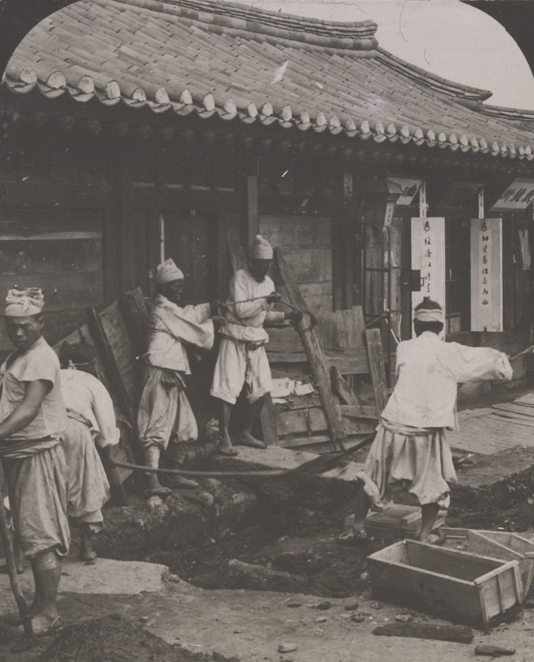 Korean labourers working on a pavement in a city street. They wear loose clothing.  The wooden building behind them has a large curved clay roof. 