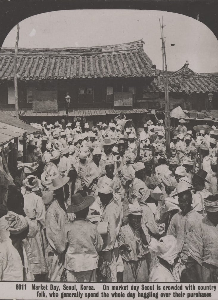 Very large crowd of people in the market place, market day, Seoul, Korea. They wear light, loose clothing and cloth hats. The wooden buildings behind them have long, tiled clay  roofs. 