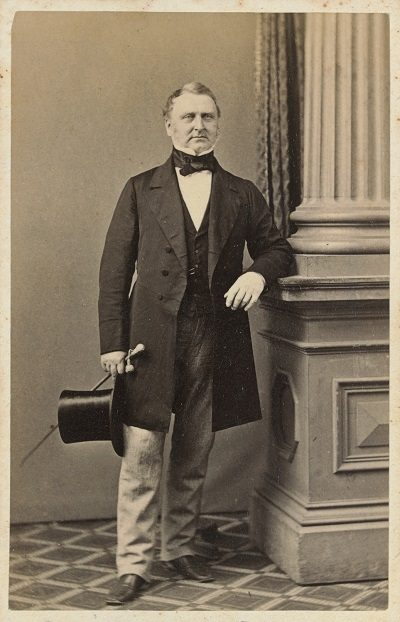 Full length portrait of Sir Redmond Barry, standing, left elbow resting on a pillar, right hand holding a hat and cane, wearing a suit with long coat and necktie.
