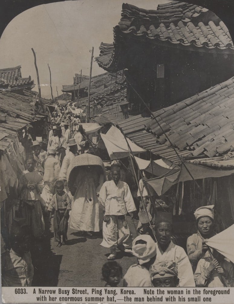 Crowds of people in narrow street, Ping Yang, korea. One woman wears an enormous summer hat and one man carries a fan.  The roofs are curved and made of clay tiles.