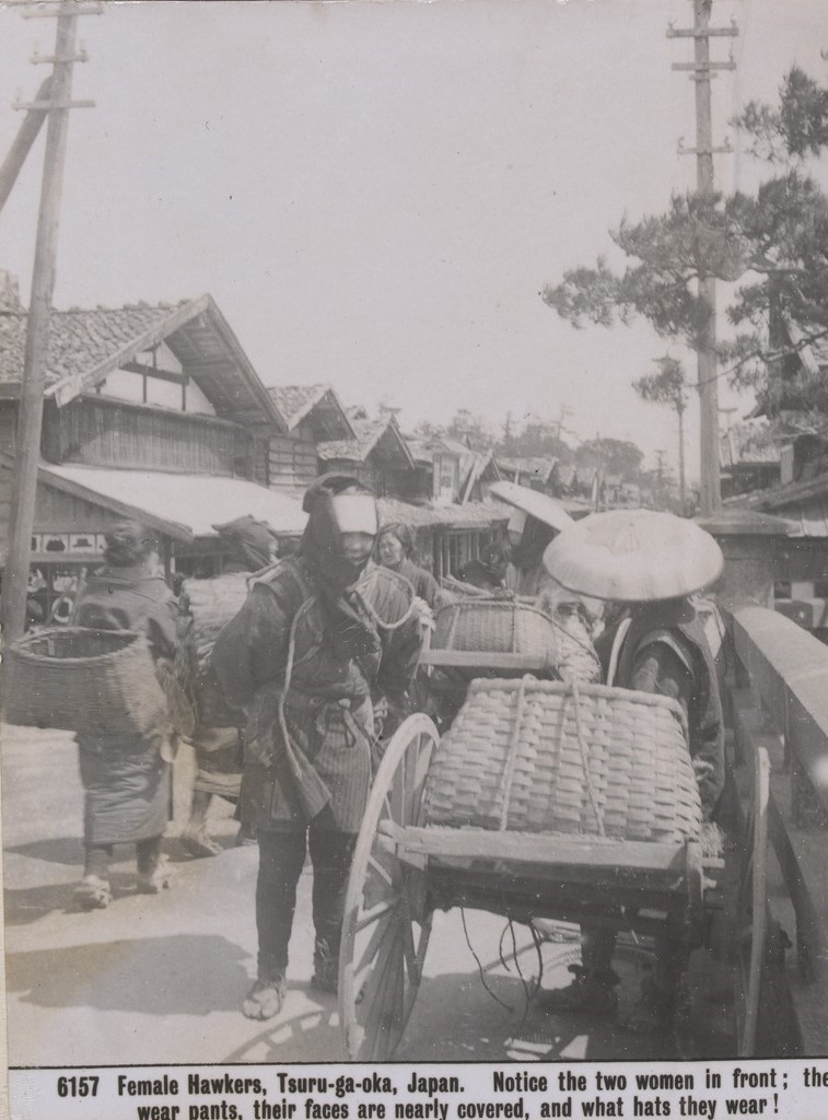 Women selling goods at the market. Some women  use large cane baskets on large wheels. One woman carries a large basket on her back.  The women in front wear trousers, long jackets and hats. Houses with gable roofs line the street. 