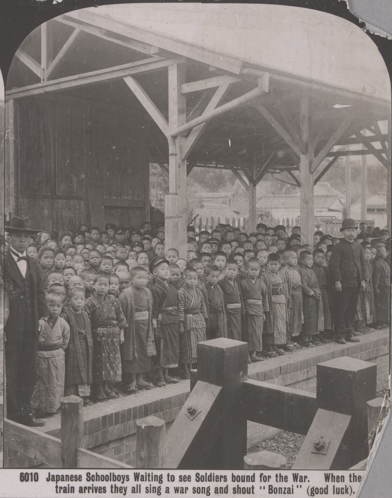 crowd  small Japanese schoolboys wearing traditional Japanese clothing wait to say for soldiers to arrive at railway station. The soldiers will go off to Russo-Japanese war, 1905. Two men wearing suits  wait with them. 