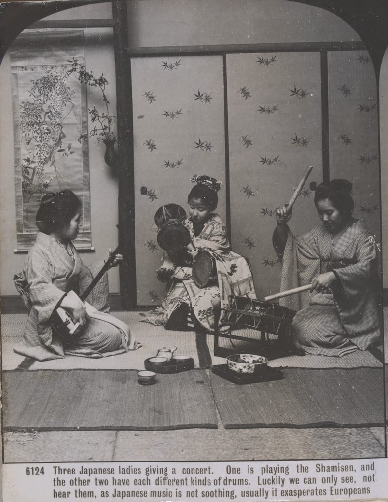 Three Japanese women wearing kimonos, seated on a mat, playing musical instruments.