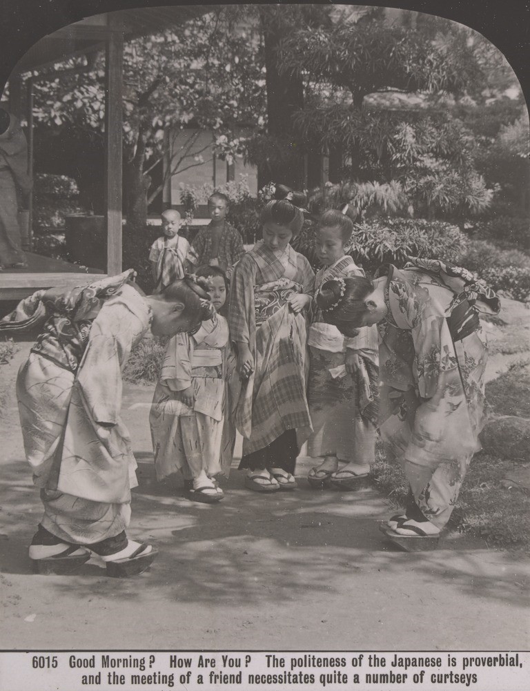 Two Japanese women wearing colourful  kimonos curtsey deeply to each other. Two other Japanese women and a little girl watch them. All the women have their hair pulled up with a bun on top.  