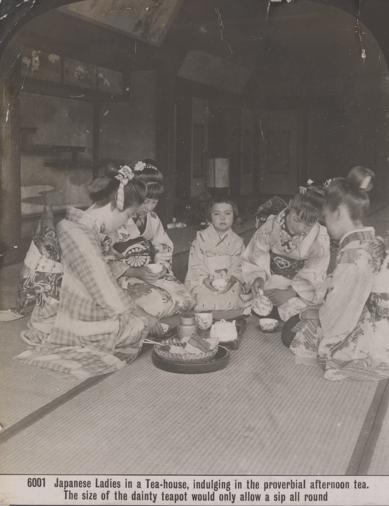 Japanese women and a little Japanese girl sitting on a mat in a tea-house. They hold tea cups while one woman pours tea from a teapot.    