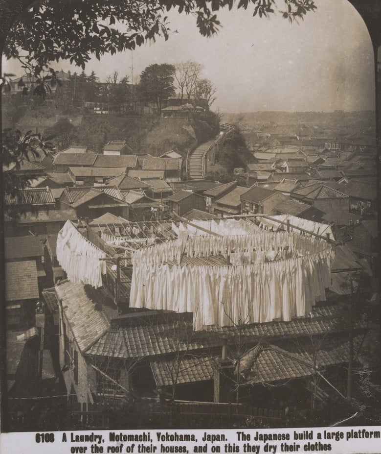 Scene showing a city of roof tops with  houses and trees on a hill. In the foreground is a large clothes line on a roof top. It is full of white cotton clothing. 