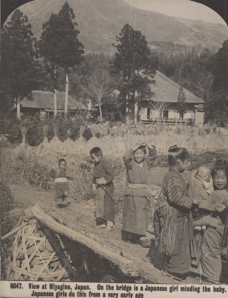Six small Japanese children wearing kimonos stand on a bridge.  One little girl holds a baby. Mountains, trees, two houses and a  field  stand behind them.  The children smile. A small boy runs.  They look very happy. 