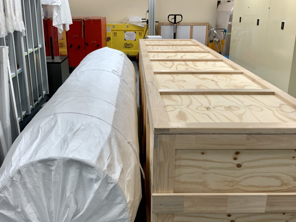 A large cylindrical object wrapped in white material rests on the floor, next to a large wooden crate. 