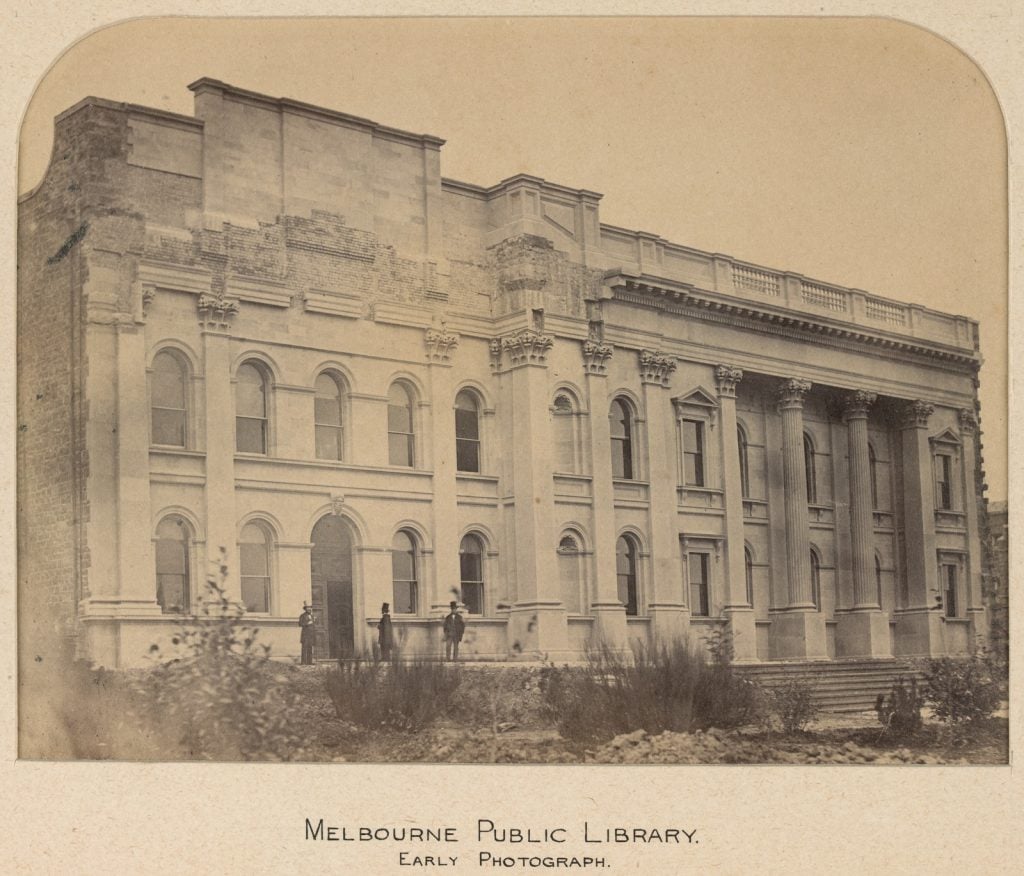 Southern frontage of Swanston Street entrance of Melbourne Public Library with garden in foreground, three men in suits and top hat standing at entry. Circa 1860.