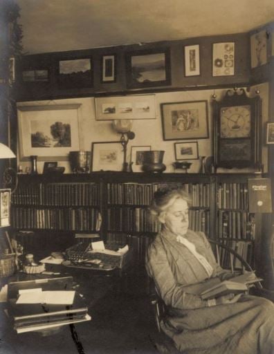Sepia-toned photographic portrait of a woman siting in an armchair reading a book, in a room full of bookcases with artworks on the walls. 