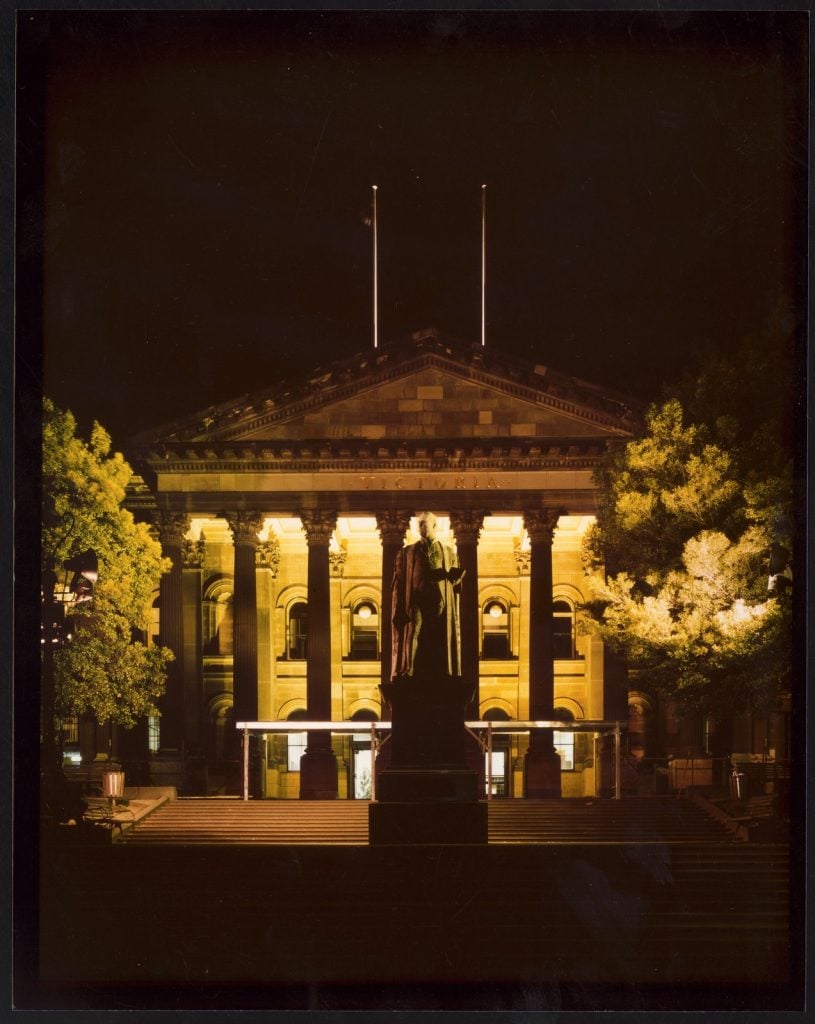 Colour photo of the State Library Victoria at night. The staue of Redmond Barry is on the steps in the foreground and the library's portico is floodlit against a dark background.