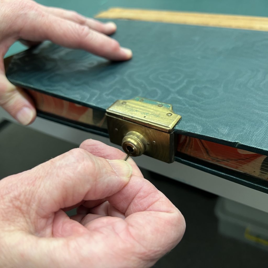 Two hands using a tool to unpick a metal lock fastened to the side of a book. 