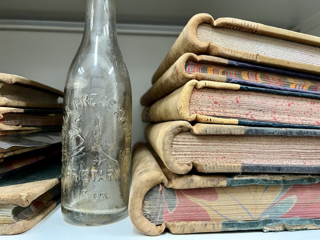 A stack of ledgers and an empty glass bottle on a shelf.