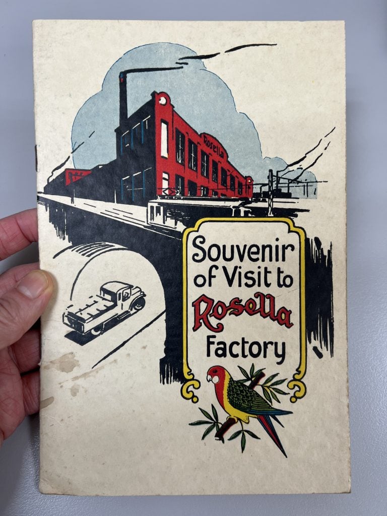 Hand holding a booklet depicting a colour illustration of the Rosella Preserving Company bird logo, factory exterior and packed truck. 