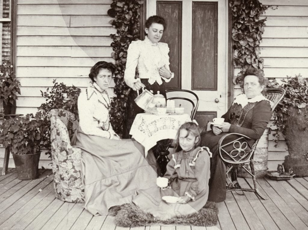 wo women sitting at a small table on verandah of a weatherboard house, another woman standing pouring tea, a little girl seated on rug at their feet also drinking tea.