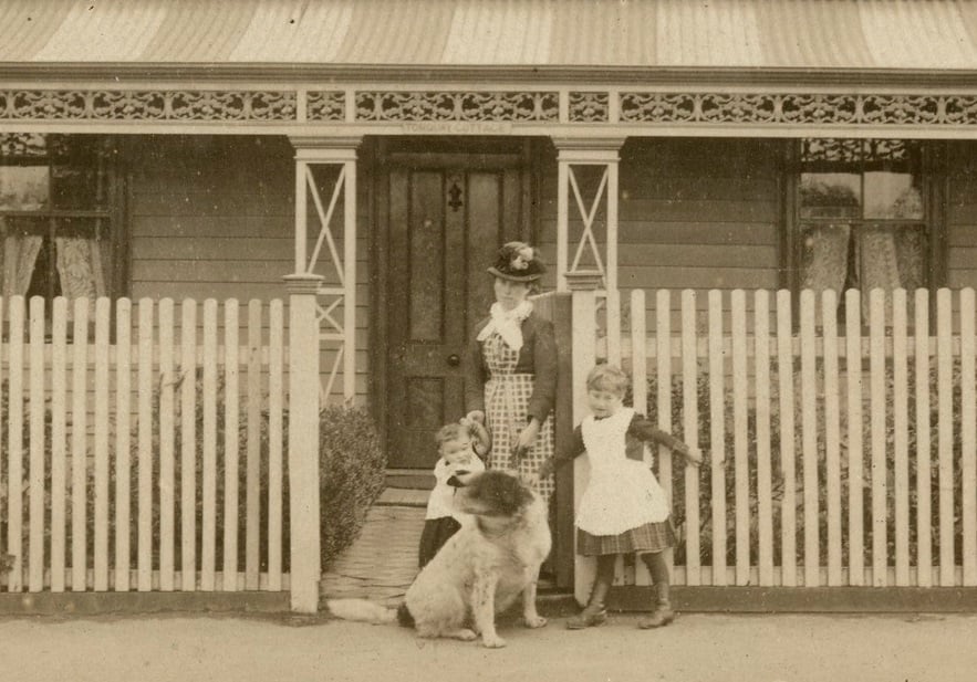 A lady wearing a hat and checked dress,  stands at the front gate of a weatherboard house, with two young children and a large dog,