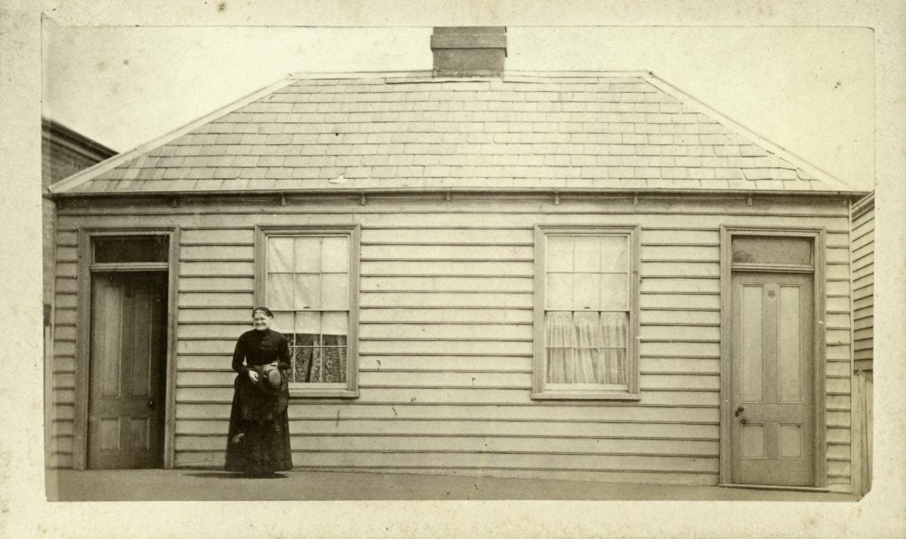 A woman stands, alone, in front of her stark weatherboard, two doors, two windows, one chimney.