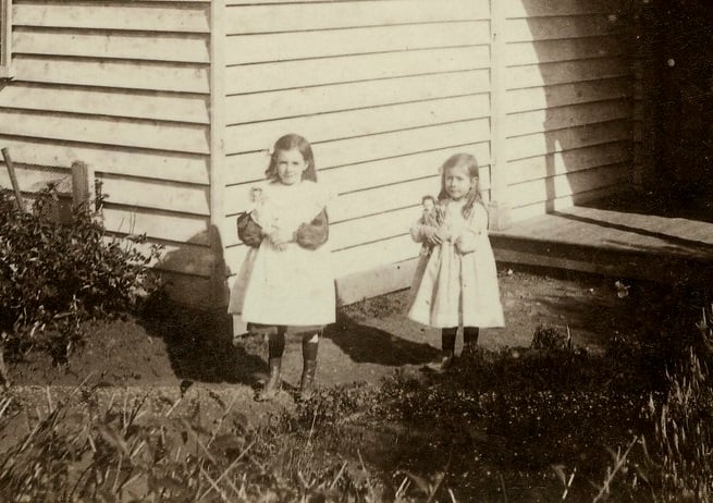 Weatherboard house with corrugated iron roof and porch, little girls standing in front wearing pinafores, both holding dolls