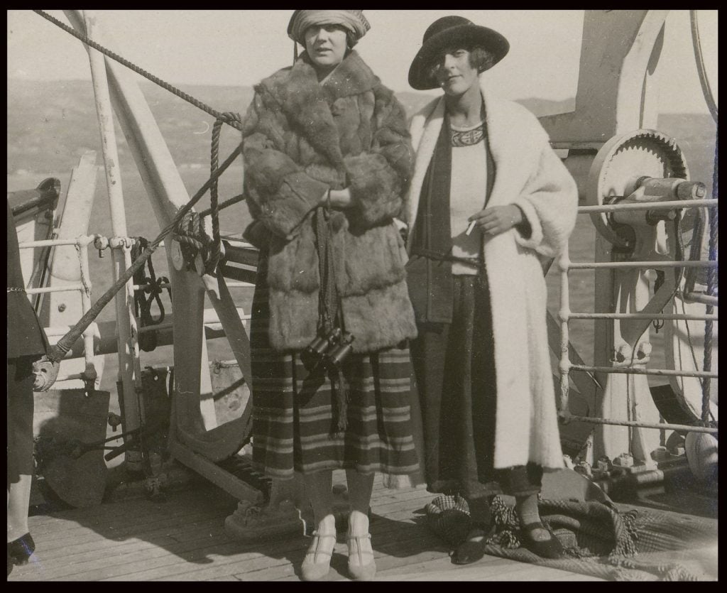 Whole-length, full face, one woman wearing three-quarter length fur coat and hat, the other a calf length coat and brimmed hat, ship's rails and ocean behind them.