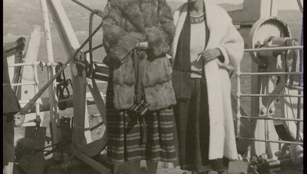 Whole-length, full face, one woman wearing three-quarter length fur coat and hat, the other a calf length coat and brimmed hat, ship's rails and ocean behind them.