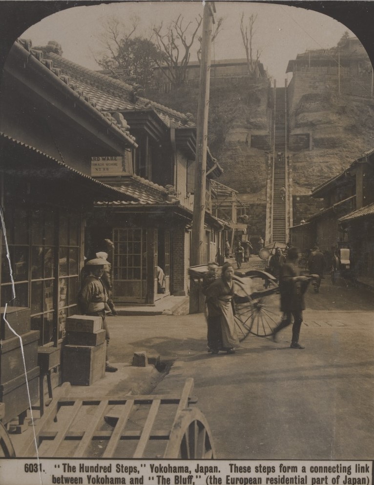 Shops in Yokohama, Japan. A woman carries a baby on her back. A man pulls a rickshaw. People stand and walk in the street. In the background are one hundred very steep steps on the side of a hill. 
