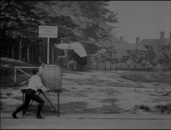 still frame of one man jumping into barrel on table. Black and white image.