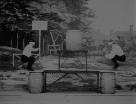 still frame of two blindfolded men jumping into barrels. Black and white image.