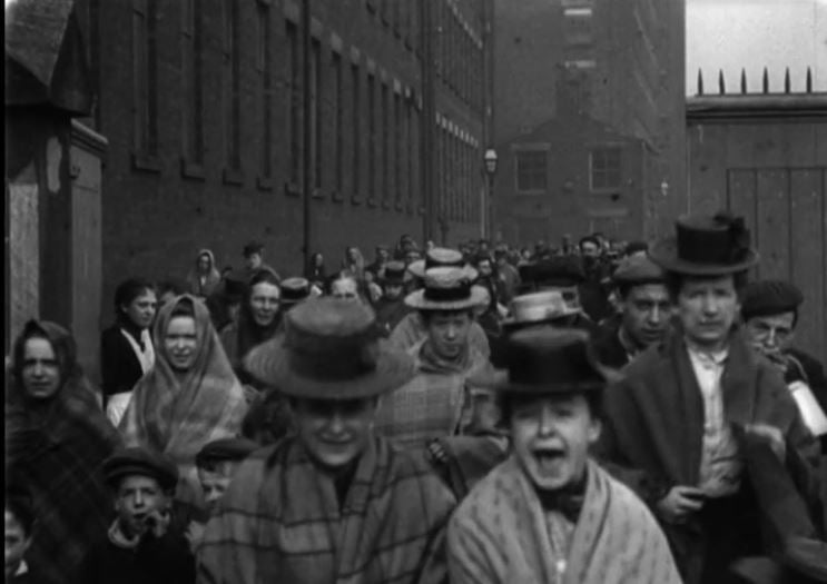 still frame of people leaving factory, one woman laughing, facing the camera. Black and white image.