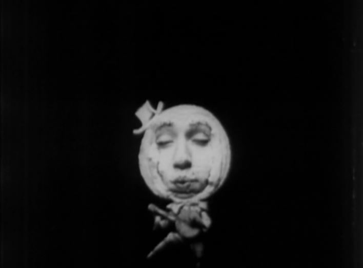 still frame of performer dressed as the moon with a little had in, with small body playing a banjo. Black and white image.