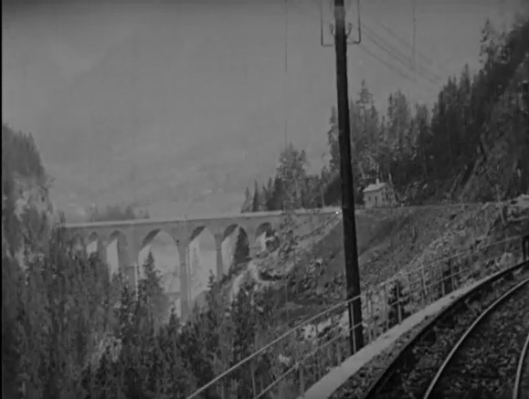 Still frame of view from the front of a train in the mountains, bridge in the background. Black and white image.