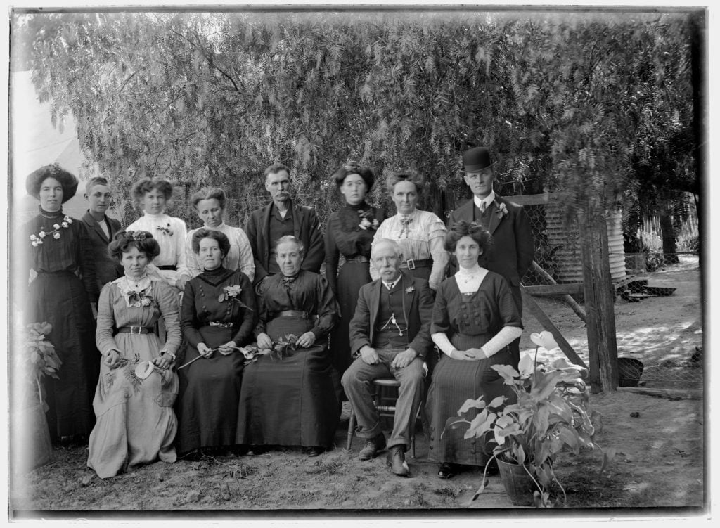 Black and white photograph of group portrait standing and seated with a large pot of foliage to the right as decoration, under a peppercorn tree